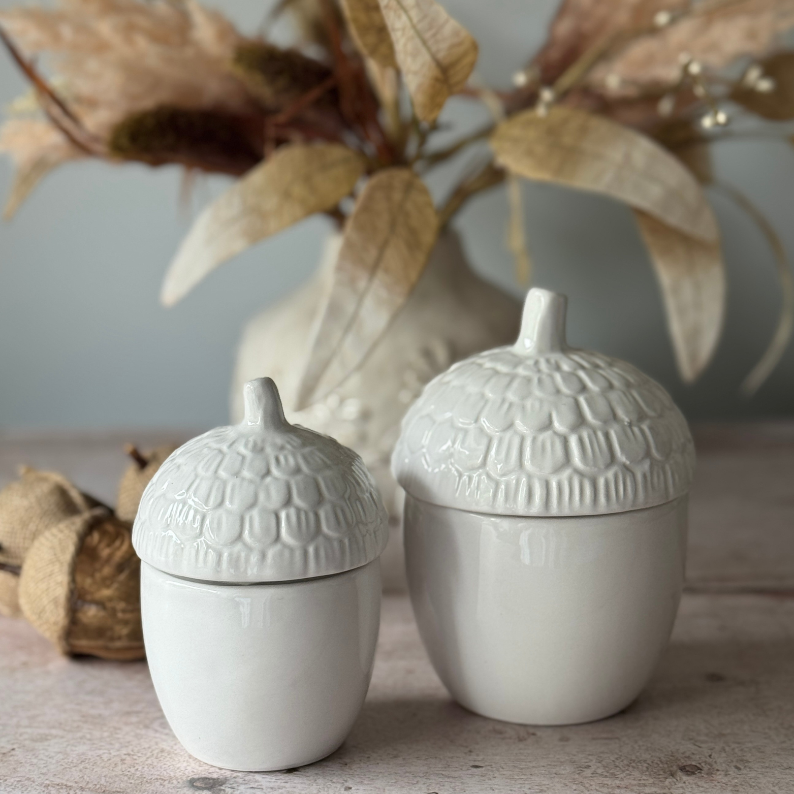 Two white textured ceramic acorn-shaped containers on a wooden surface with dried plants in the background.