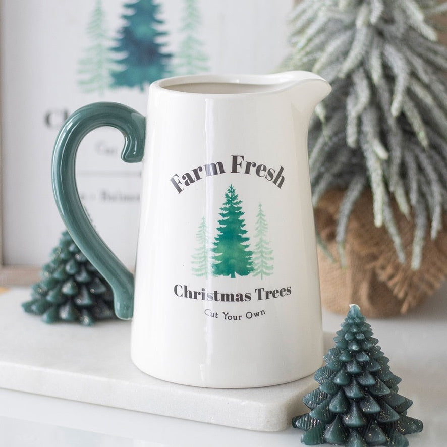 White ceramic pitcher with 'Farm Fresh Christmas Trees Cut Your Own' text and tree graphics, surrounded by decorative trees.