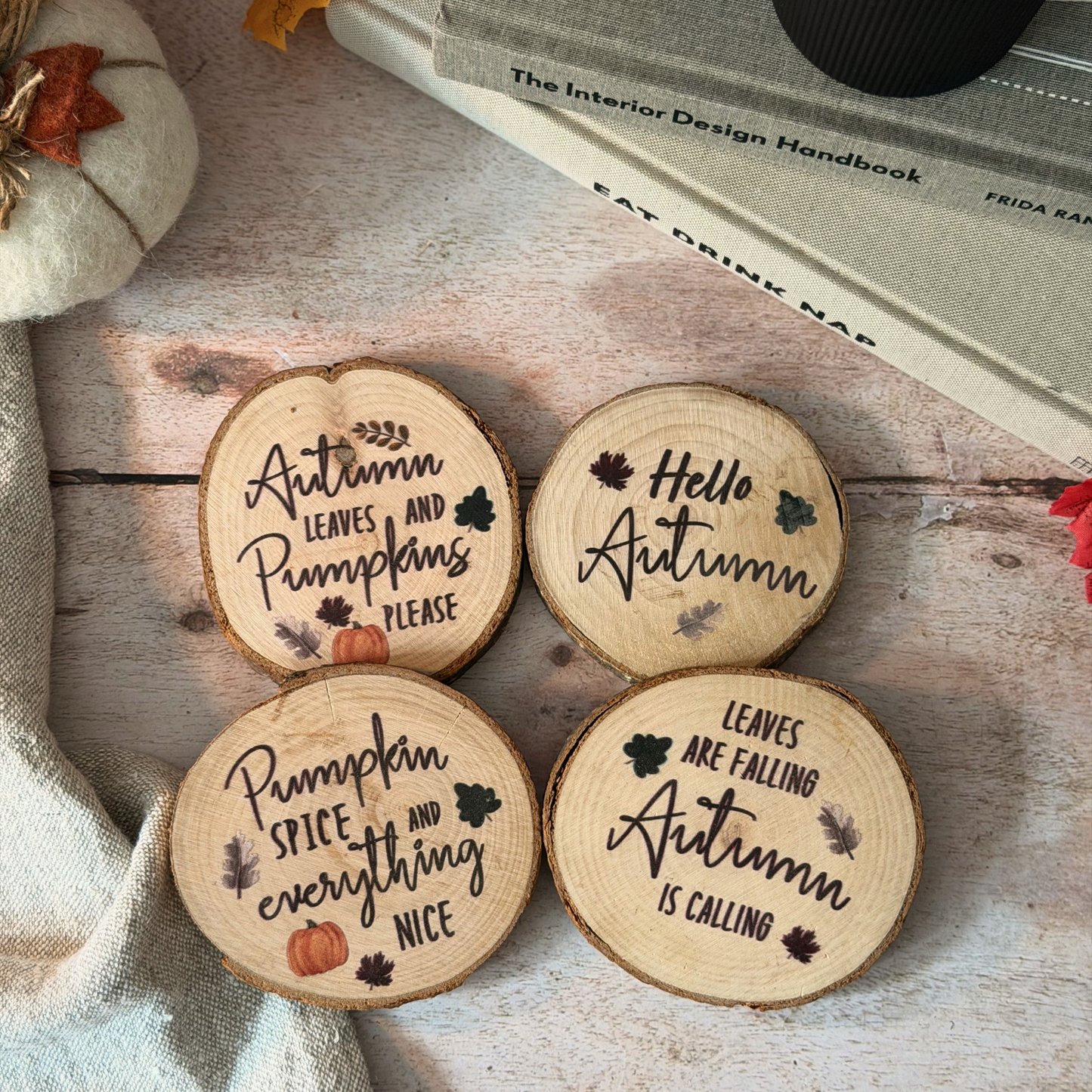 Four round wooden coasters with autumn-themed engraved quotes, arranged on a rustic wooden surface with a small pumpkin and books in the background.