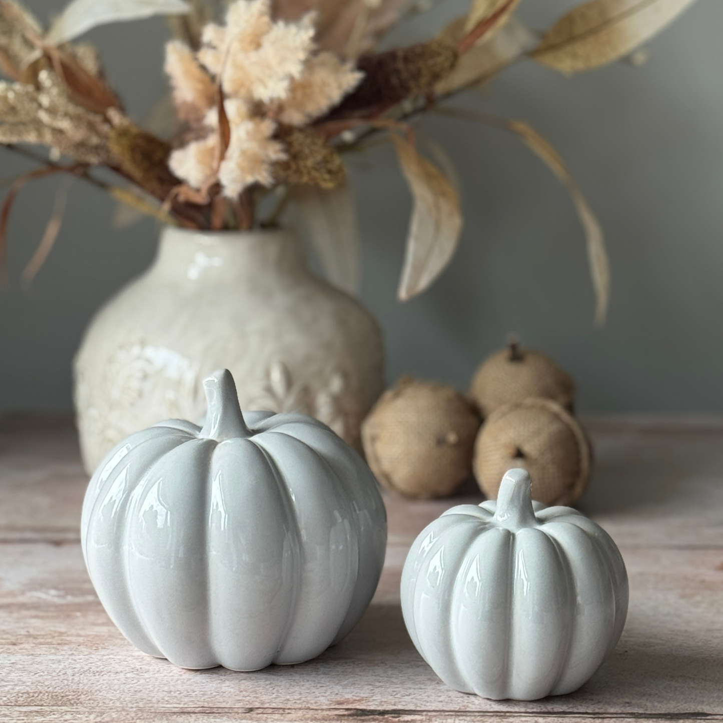 Two grey ceramic pumpkins on a wooden surface with a vase and acorns in the background.