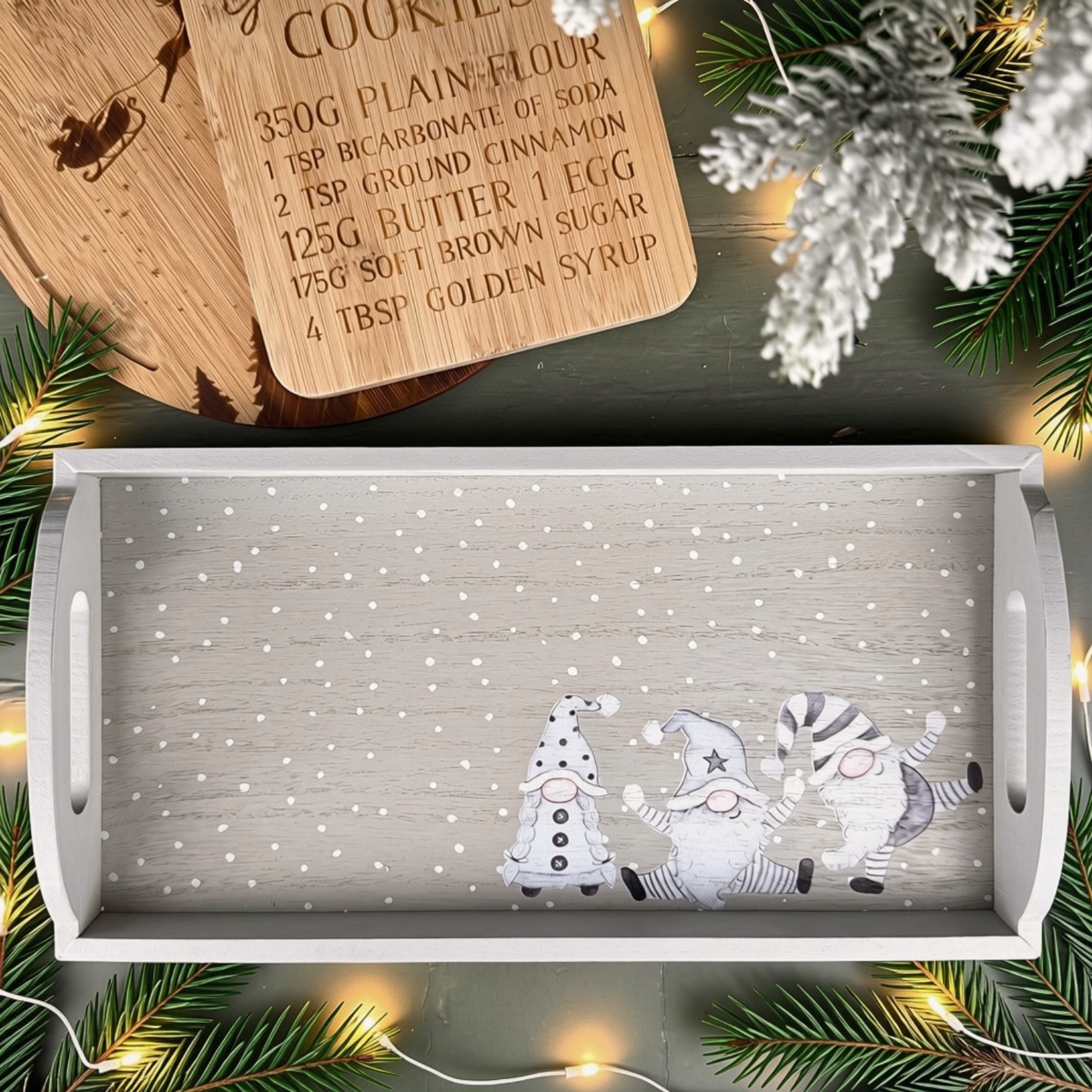 A rectangular wooden serving tray with a snowy white-grey grained surface and a printed illustration of three gnomes at the bottom, styled on a dark tabletop with pine branches, fairy lights, a wooden recipe card, and white snow-dusted evergreen accents.