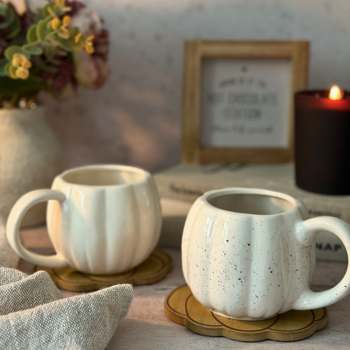 Two cream-colored ceramic mugs with a ribbed pumpkin-like texture, one solid cream and the other speckled cream, sitting on wooden coasters on a table with a candle, books, and a framed print in the background.