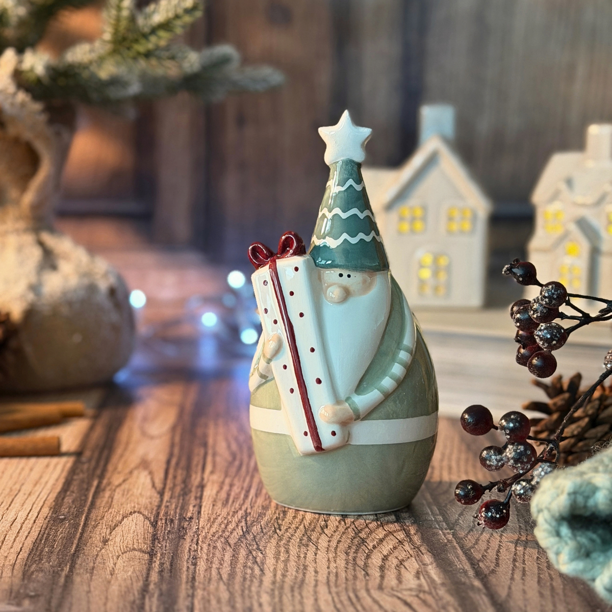 A ceramic Christmas santa figurine holding a wrapped gift, styled on a wooden tabletop with blurred holiday decor and miniature houses in the background.
