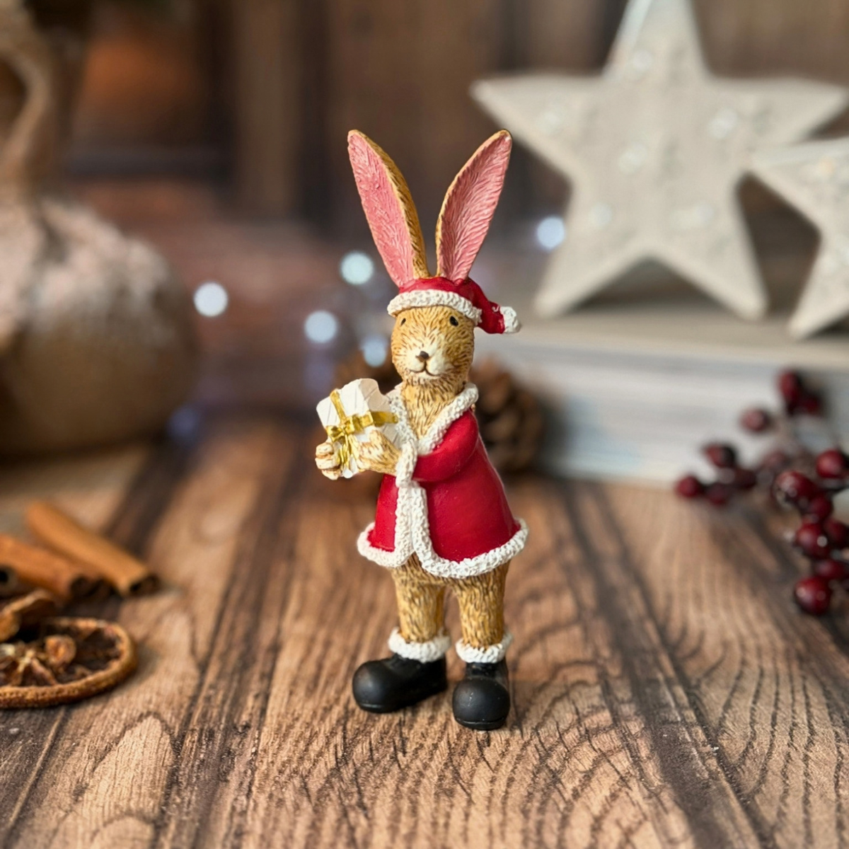 A small decorative rabbit figurine dressed in a red Santa-style coat and hat, holding a wrapped gift, displayed on a wooden surface with holiday props in the background.