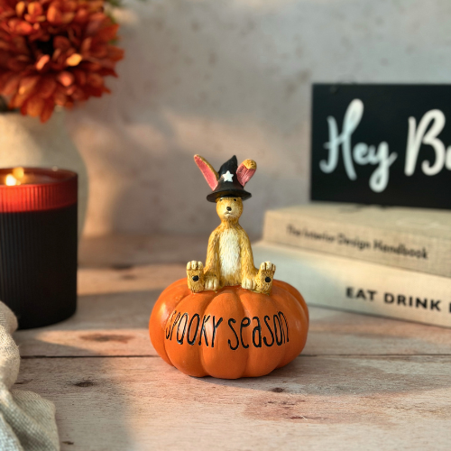 A decorative figurine of a golden bunny wearing a black witch hat sitting on an orange pumpkin with the words 'SPOOKY season' written on it, styled on a wooden surface with a candle, books, and autumn flowers in the background.