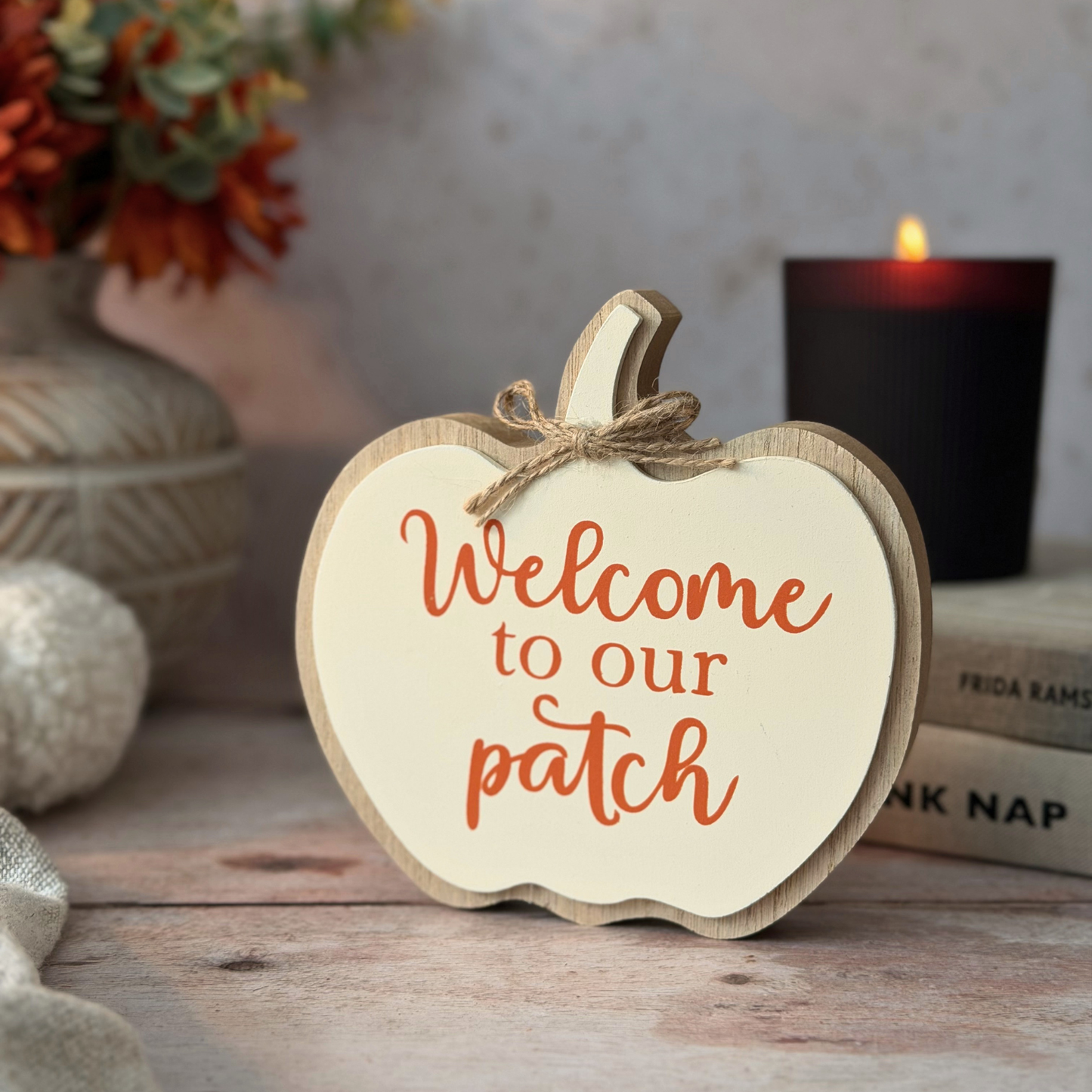 A wooden apple-shaped sign with the phrase 'Welcome to our patch' in orange script, styled on a rustic tabletop with a lit candle, books, and autumn decor in the background.