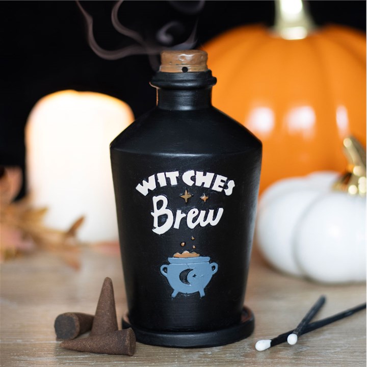 A black incense burner designed to look like a 'Witches Brew' potion bottle, with a label that includes the text 'WITCHES BREW' and an illustration of a witch's hat and a cauldron. The bottle is placed on a wooden surface with a pumpkin and incense cones nearby.