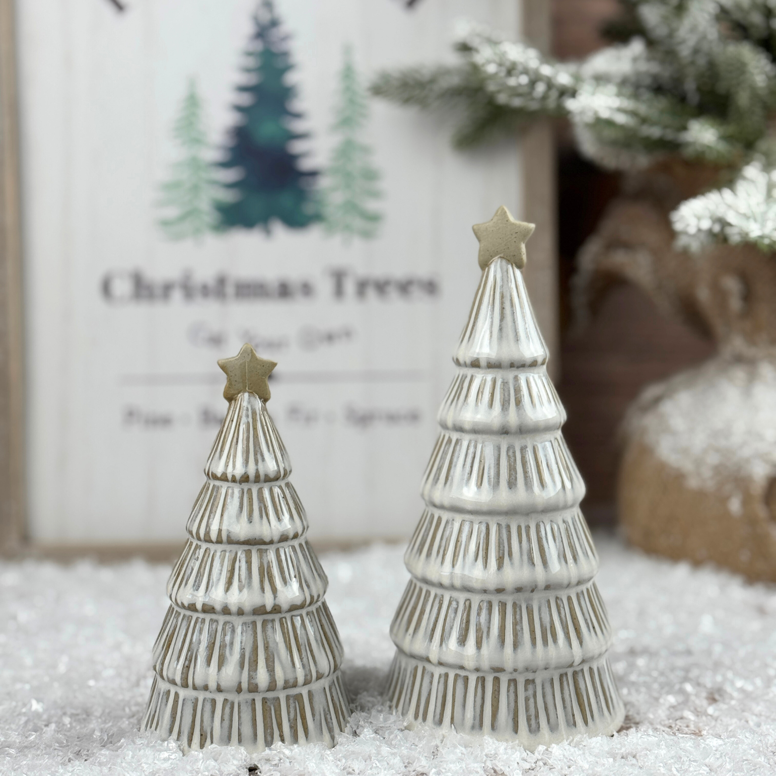 Two Christmas tree figurines with stars on a snowy surface, with a framed picture of trees in the background.