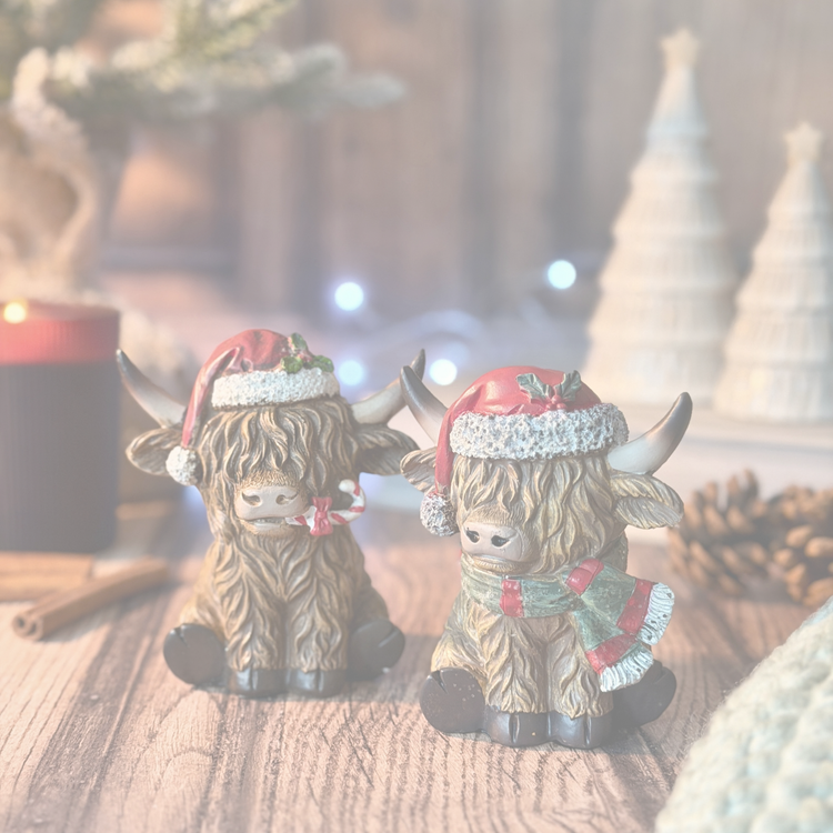Two Highland cow figurines decorated for Christmas, sitting on a wooden surface with a blurred festive background containing candles, pinecones, and holiday lights.