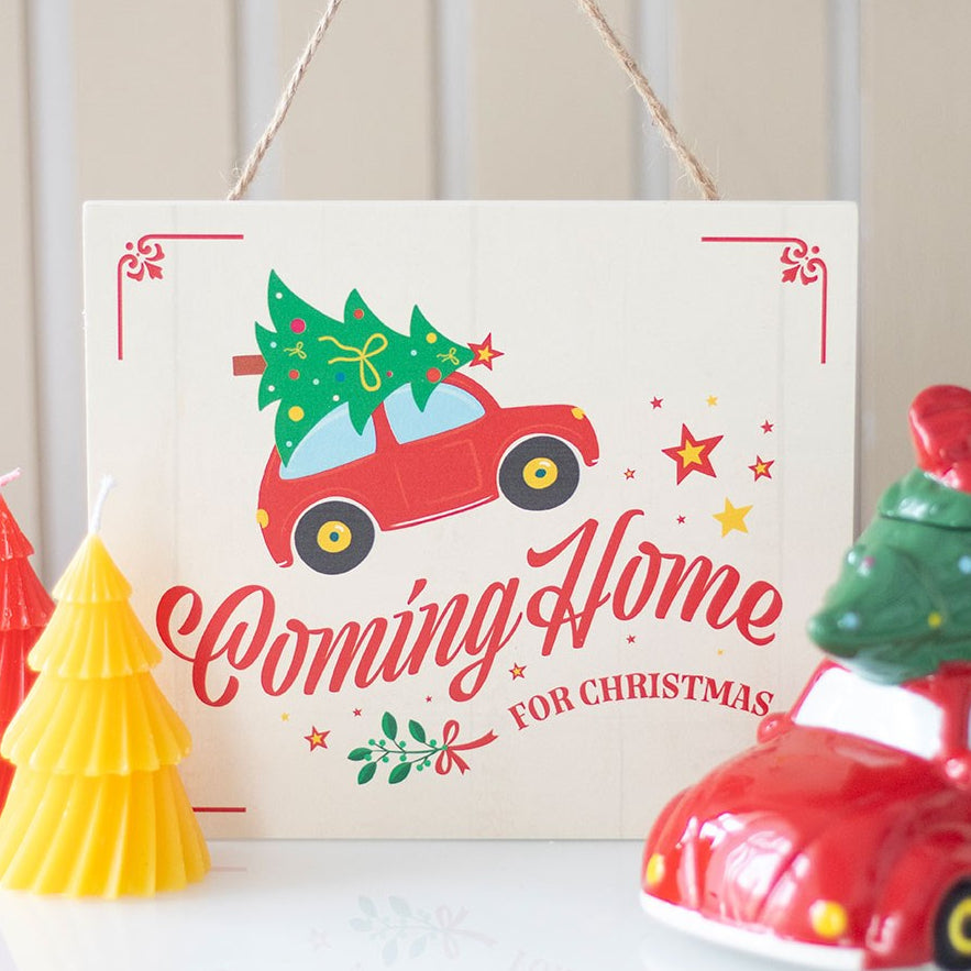 Decorative sign with a red car carrying a Christmas tree and candles on a white surface.