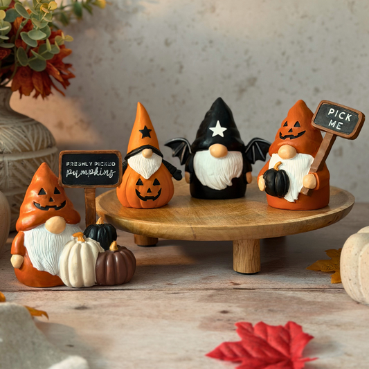 Four Halloween gnome figurines displayed on a round wooden stand, with two smaller pumpkins in front and small chalkboard signs reading 'PICK ME' and 'FRESHLY PICKED PUMPKINS'. The scene is styled with autumn leaves, a vase, and a textured wall.