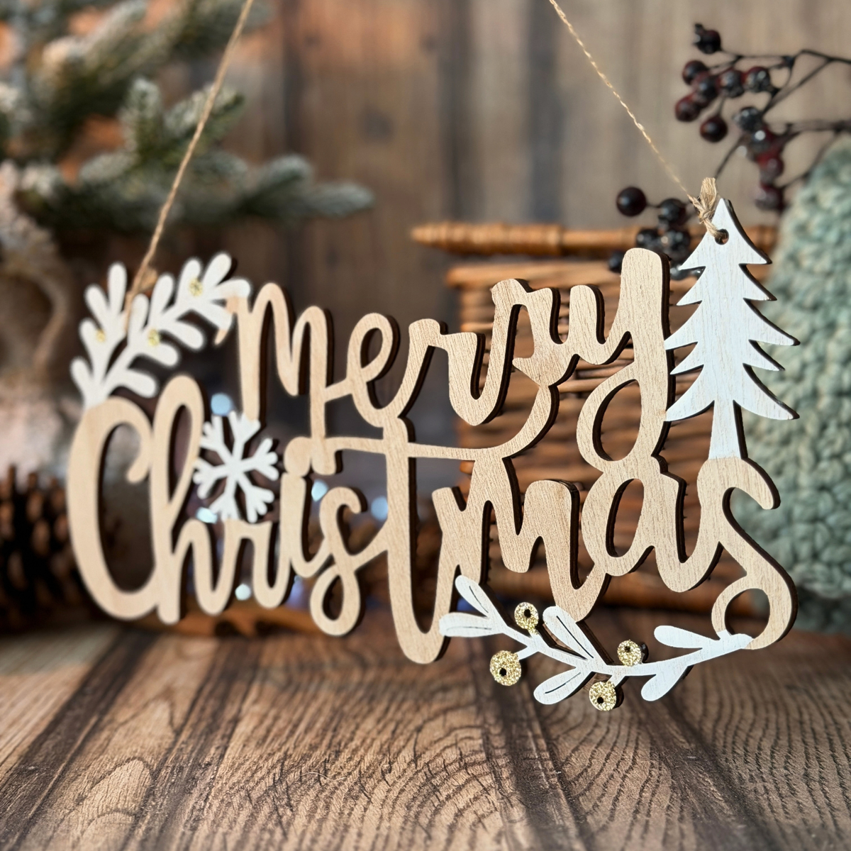 A wooden hanging Christmas sign that reads “Merry Christmas” in cursive, with white leaf and snowflake cutouts and small gold bead accents, displayed on a rustic wooden surface with holiday decor in the background.