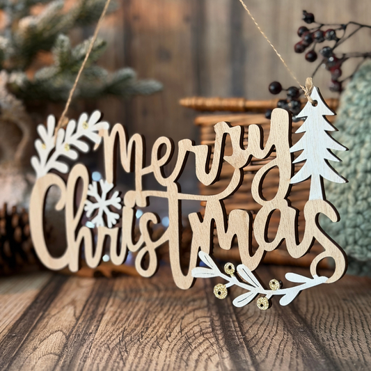 A wooden hanging Christmas sign that reads “Merry Christmas” in cursive, with white leaf and snowflake cutouts and small gold bead accents, displayed on a rustic wooden surface with holiday decor in the background.