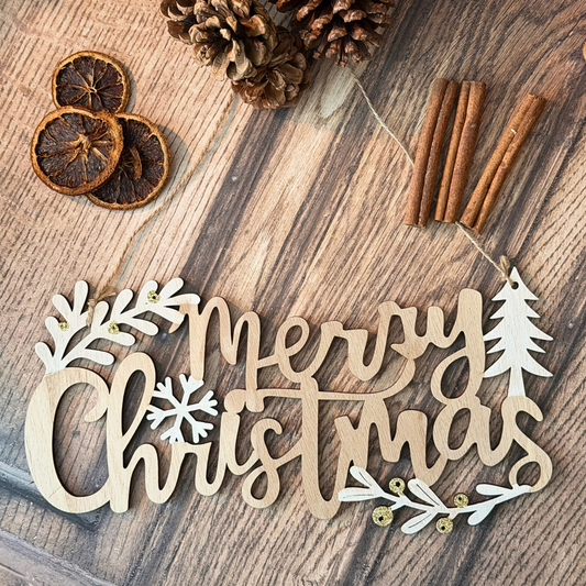 A wooden 'Merry Christmas' cutout sign with white leaf and snowflake accents, styled on a rustic wooden surface with dried orange slices, pinecones, cinnamon sticks, and small gold beads.