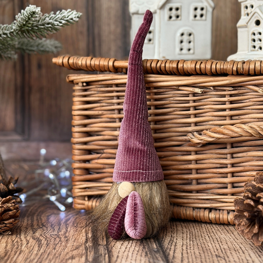 A small Christmas gonk with a tall plum corduroy hat, brown faux-fur beard, and pink nose, posed in front of a wicker basket on a rustic wooden surface.
