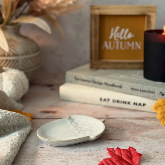 A styled autumn tabletop scene with a small off-white ceramic dish in the foreground, stacked books, a candle, dried flowers, and a framed sign reading 'Hello AUTUMN'.