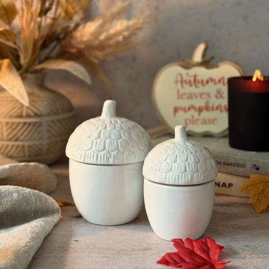Two white ceramic acorn-shaped containers with textured lids, styled on a rustic wooden surface with autumn props including a candle, a sign reading 'Autumn leaves & pumpkins please', and dried grass in a vase.