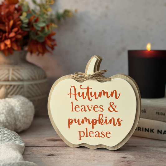 A scalloped wooden pumpkin-shaped sign with the phrase 'Autumn leaves & pumpkins please' in orange lettering, styled on a rustic table with a vase of flowers, a candle, and books in the background.