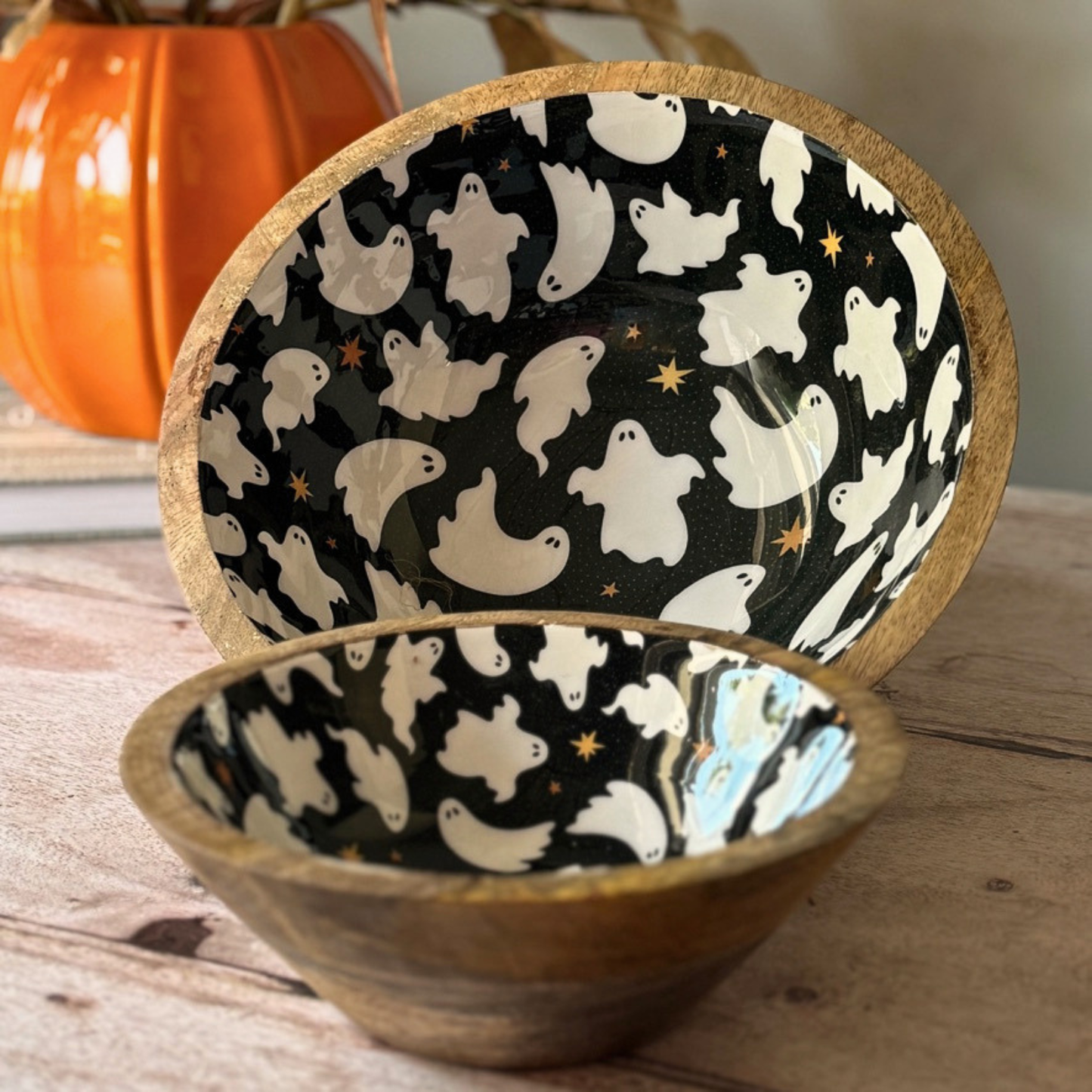 Two wooden bowls with a black and white ghost pattern on the inside, displayed on a rustic wooden table with a decorative orange pumpkin in the background.
