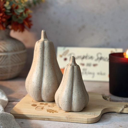 Two speckled ceramic pumpkins in different sizes displayed on a wooden cutting board, with a lit candle and fall-themed decor in the background.
