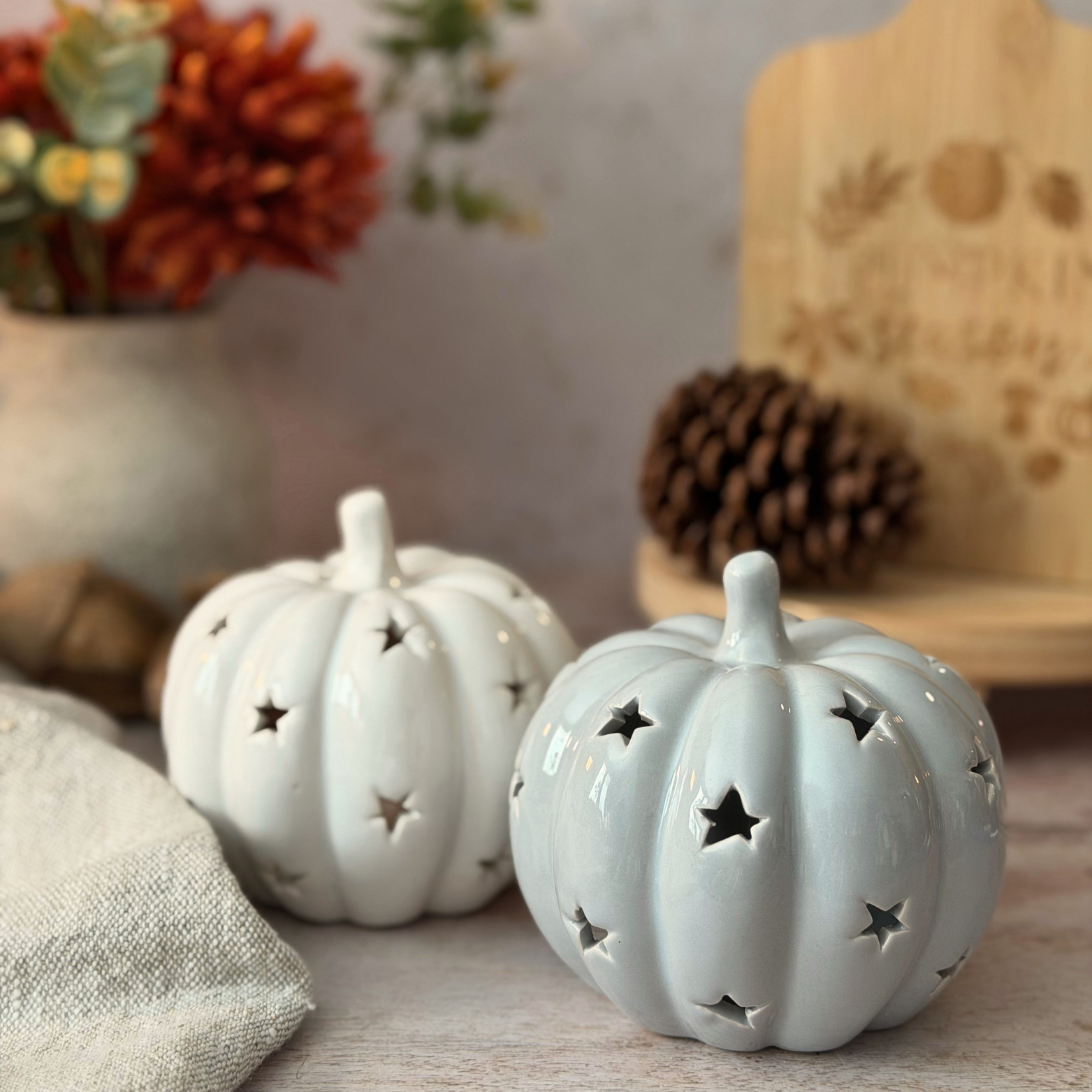 Two ceramic decorative pumpkins with star-shaped cutouts, styled on a tabletop with autumn props including a pinecone, a vase of flowers, and a wooden sign in the background.