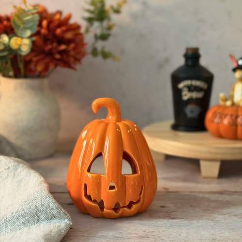 An orange ceramic jack-o'-lantern candle holder with carved facial features, styled on a rustic wooden surface with autumn-themed props in the background.