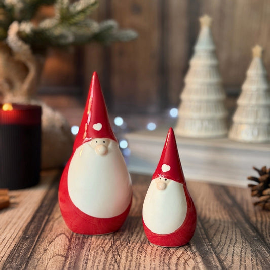 Two small red and white gnome figurines standing on a rustic wooden surface, with blurred Christmas decorations in the background including white tree ornaments and a candle.
