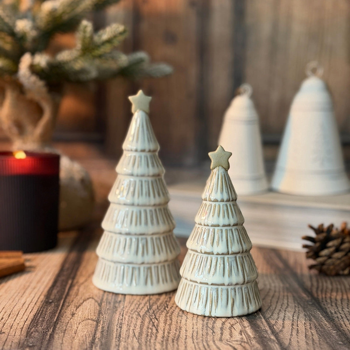 Two small white ceramic Christmas tree ornaments with textured tiered layers and star toppers, displayed on a wooden surface with a blurred holiday background.