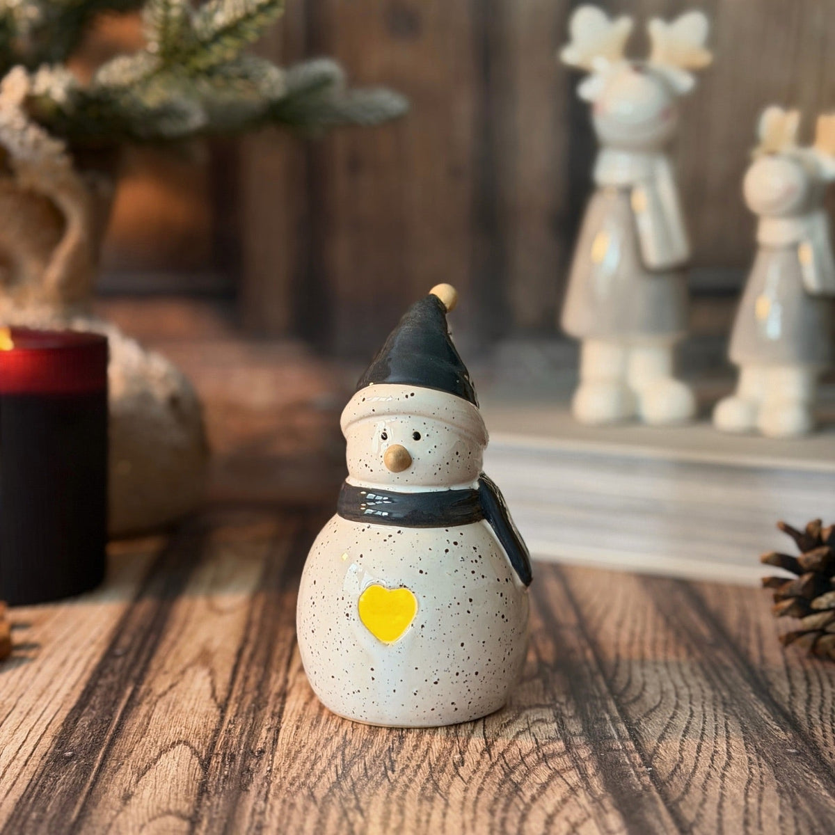 A ceramic snowman figurine with a speckled cream body, black hat and scarf, and a glowing yellow heart, displayed on a wooden surface with a candle and blurred holiday decorations in the background.