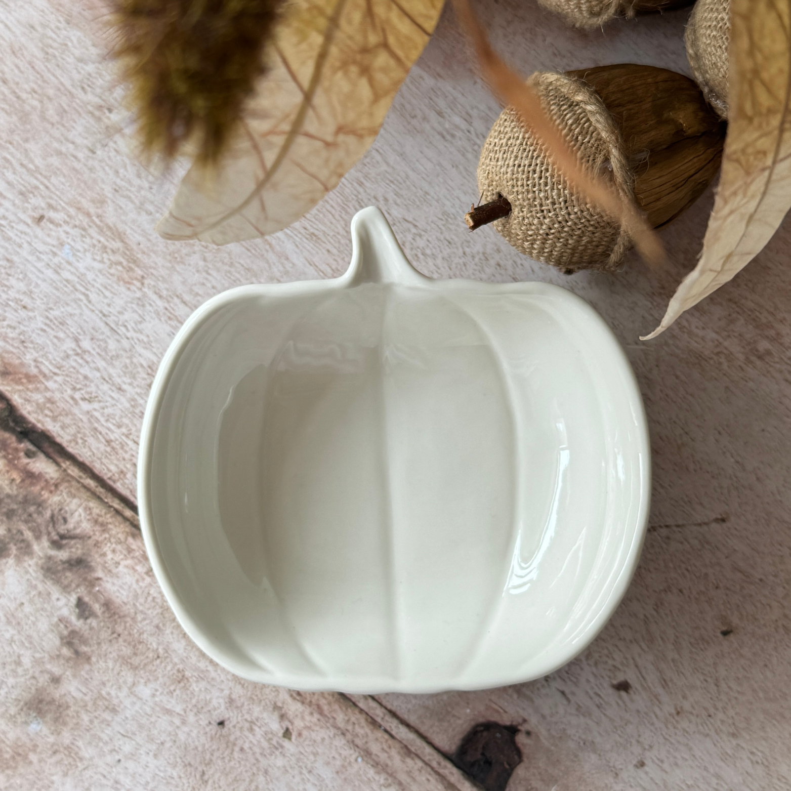 White ceramic pumpkin-shaped bowl on a wooden surface with leaves and acorns.