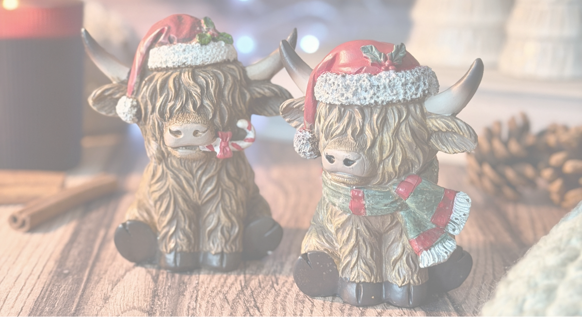 Two Highland cow figurines decorated for Christmas, sitting on a wooden surface with a blurred festive background containing candles, pinecones, and holiday lights.