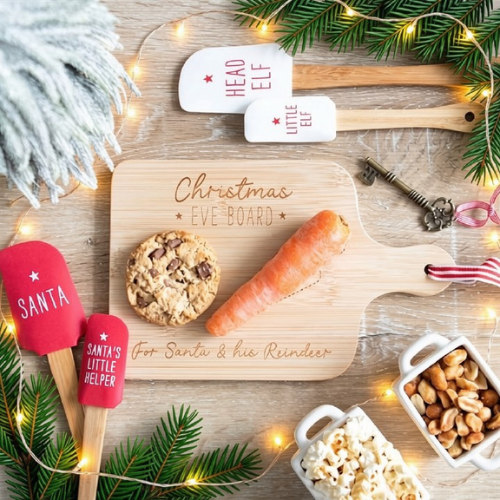 A styled flat-lay of Christmas Eve themed wooden cutting boards and spatulas on a rustic wood surface with fairy lights, pine branches, a cookie, a carrot, popcorn, peanuts, and a key. The central product is a light natural wood cutting board engraved with 'Christmas Eve Board For Santa & his Reindeer'.