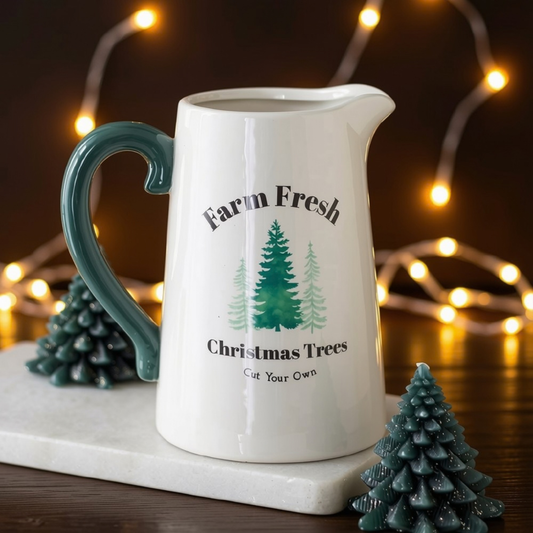 A white ceramic pitcher with a dark teal handle and printed green Christmas tree graphic, reading 'Farm Fresh Christmas Trees Cut Your Own.' It is styled on a white marble board with small teal tree figurines and warm string lights in the background.