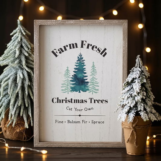 A framed rustic Christmas sign with the text 'Farm Fresh Christmas Trees' and illustrations of three pine trees, styled on a dark wooden surface with string lights and small potted frosted trees.