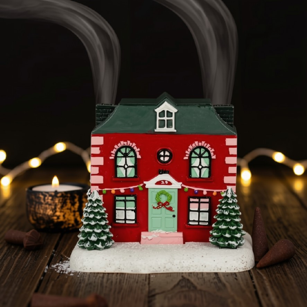 A small red Christmas house incense burner with a green roof, snowy base, two decorated trees, and glowing string lights in the background on a wooden surface.