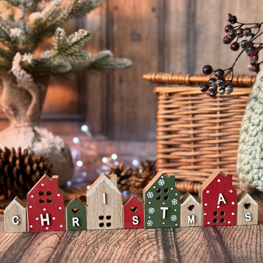 A row of small wooden house-shaped sign spelling CHRISTMAS on a rustic wooden surface, with a wicker basket, pinecones, and soft holiday decor blurred in the background.
