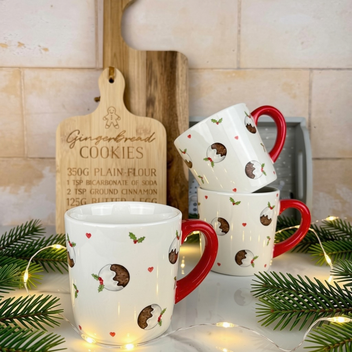 Three white ceramic mugs with red handles and a Christmas pudding and holly print, styled on a countertop with pine branches, fairy lights and a wooden cutting board.