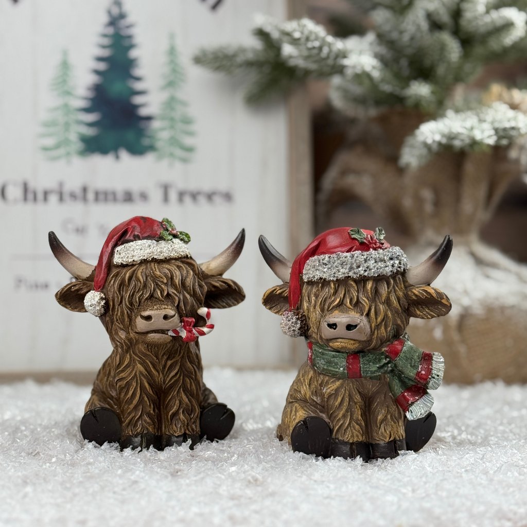 Two decorative highland cows wearing Santa hats on a snowy surface with Christmas trees in the background.