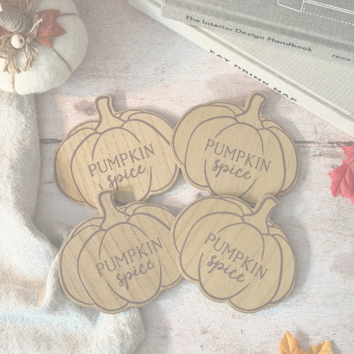 Four pumpkin-shaped wooden coasters engraved with 'Pumpkin Spice' in a warm autumnal setting, surrounded by a small decorative pumpkin, fabric, and books.