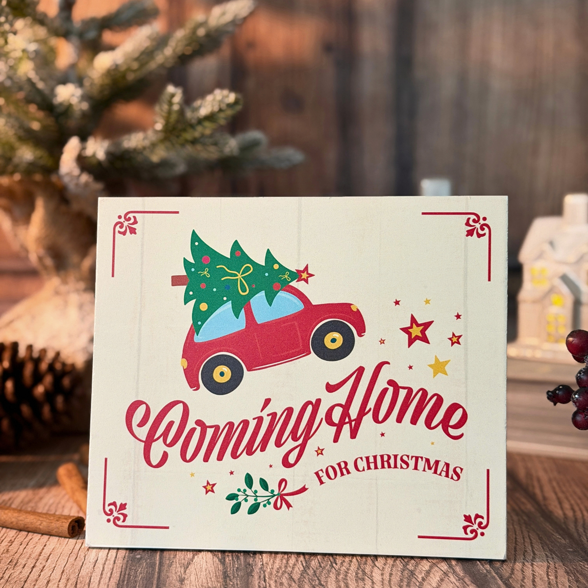 A Christmas sign with a red car carrying a green Christmas tree, with the words 'Coming Home FOR CHRISTMAS' in red script, displayed on a wooden surface with holiday decorations in the background.