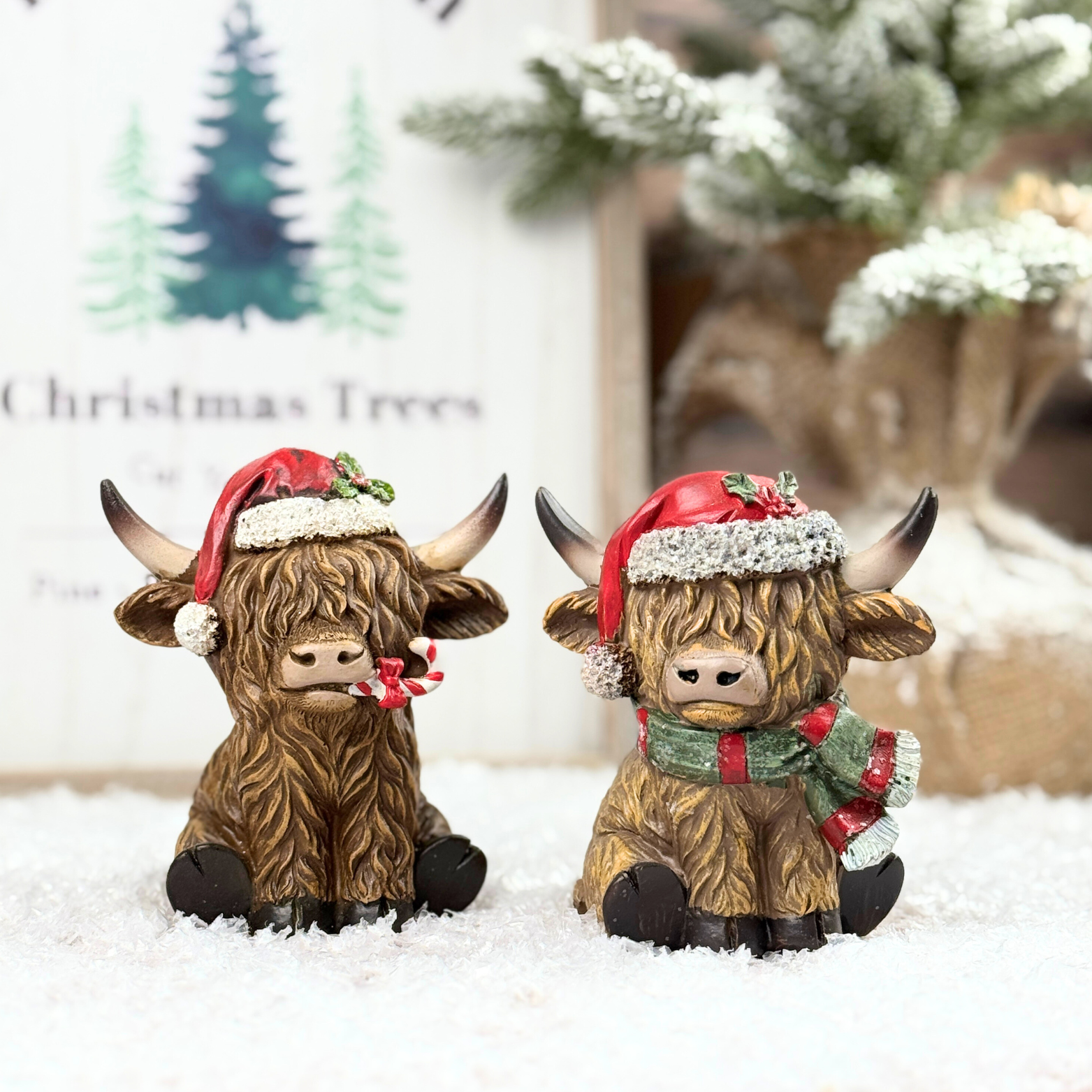 Two decorative Highland cow figurines wearing Santa hats and scarves on a snowy background.