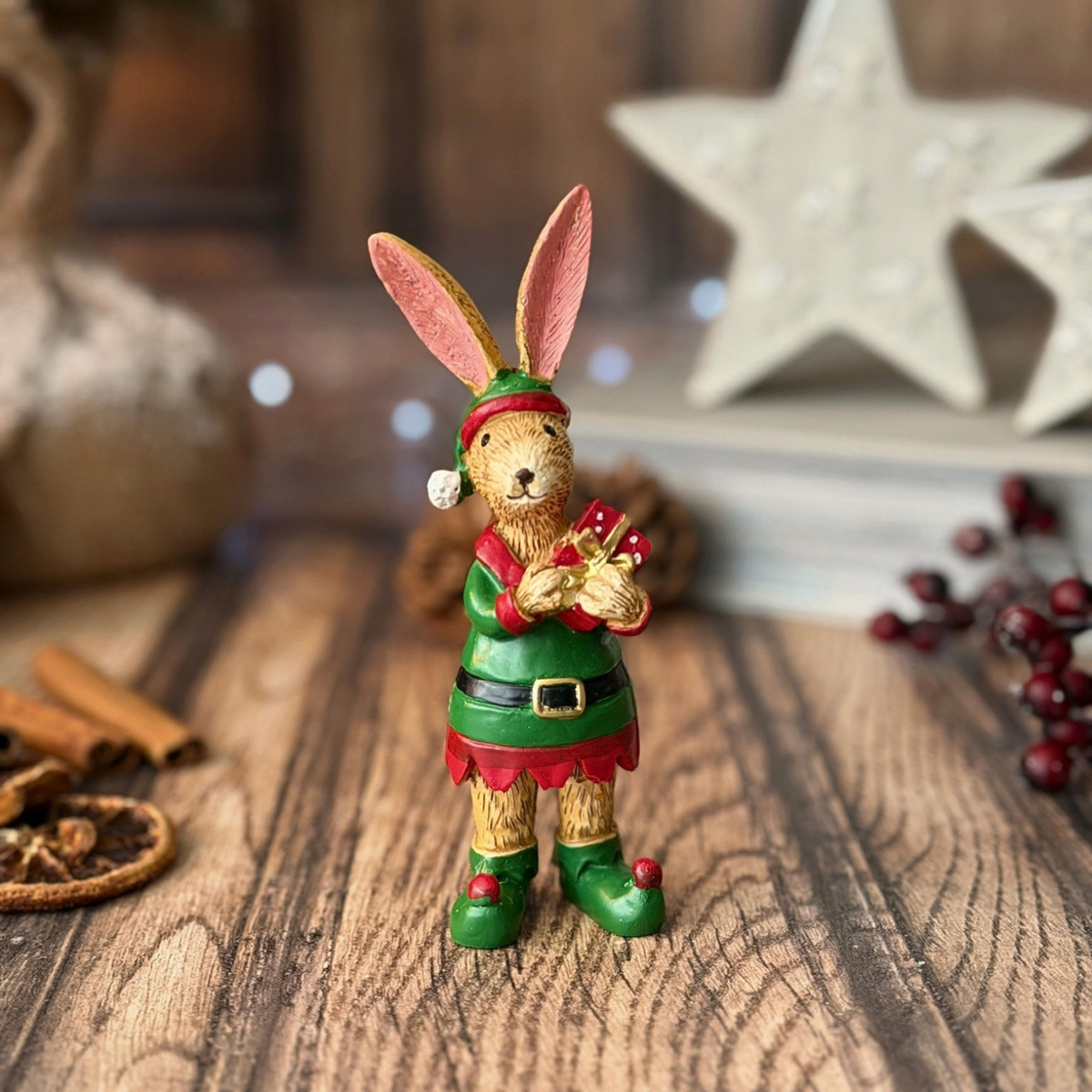 A small decorative rabbit figurine dressed as an elf stands on a rustic wooden surface. The rabbit has pink-tinted ears, a green and red outfit with a black belt, and green shoes, holding a red gift box.