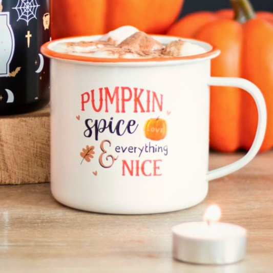 A white enamel mug with an orange rim and handle, printed with the phrase 'PUMPKIN spice & everything NICE' in orange and navy lettering, filled with a hot cocoa drink and topped with whipped cream and a marshmallow. The mug is set on a wooden surface with pumpkins in the background and a small tea light candle in the foreground.