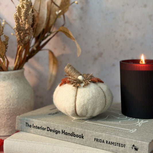 A small cream-colored felt pumpkin decoration with a burlap stem and orange leaves sits on top of two books, with a lit black candle and a vase of dried flowers in the background.