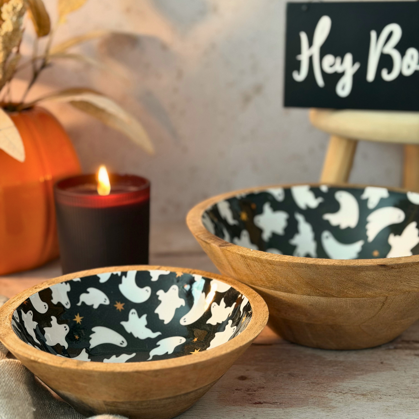 Two wooden bowls with a black and white ghost pattern on the inside, styled on a tabletop with a lit candle, an orange pumpkin, and a sign in the background.