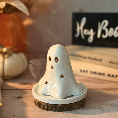 A white ceramic ghost-shaped incense holder with star and eye cutouts, sitting on a rustic wood slice base with smoke rising, styled on a wooden table with autumn props.