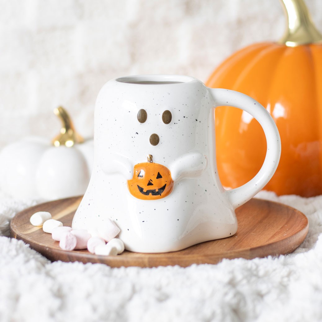 White ghost-shaped mug with a pumpkin design on a wooden coaster, surrounded by pumpkins and marshmallows.