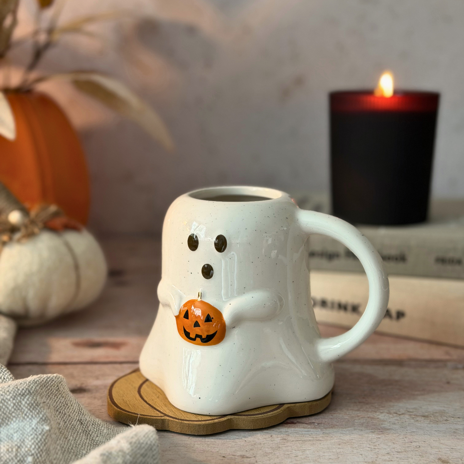 A white ceramic ghost-shaped mug with a small orange jack-o'-lantern accent, styled on a wooden surface with a lit black candle and autumn decor in the background.