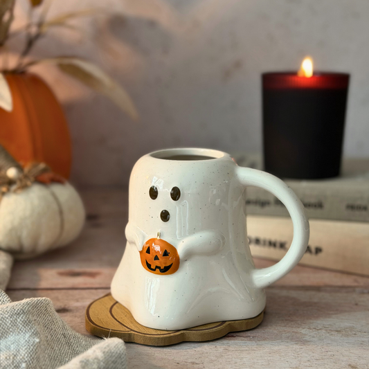 A white ceramic ghost-shaped mug with a small orange jack-o'-lantern accent, styled on a wooden surface with a lit black candle and autumn decor in the background.