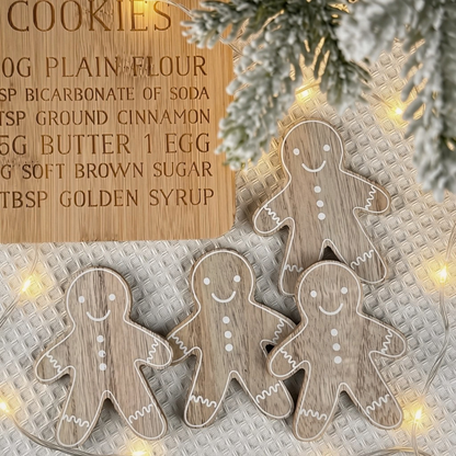 Four wooden gingerbread man coasters with white painted outlines and buttons are arranged on a textured silver surface with warm fairy lights. A wooden recipe card reading 'Cookies' is partially visible in the background, along with frosted greenery and string lights.